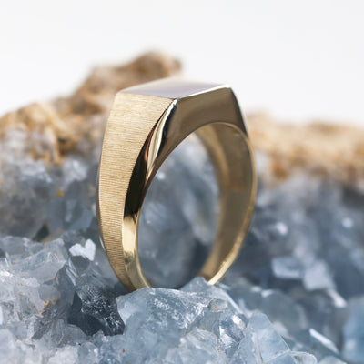 Gold signet ring on rock, close-up view. Premium metal options: sterling silver, gold, platinum. Customizable with gemstones. Dimensions and finishes detailed.