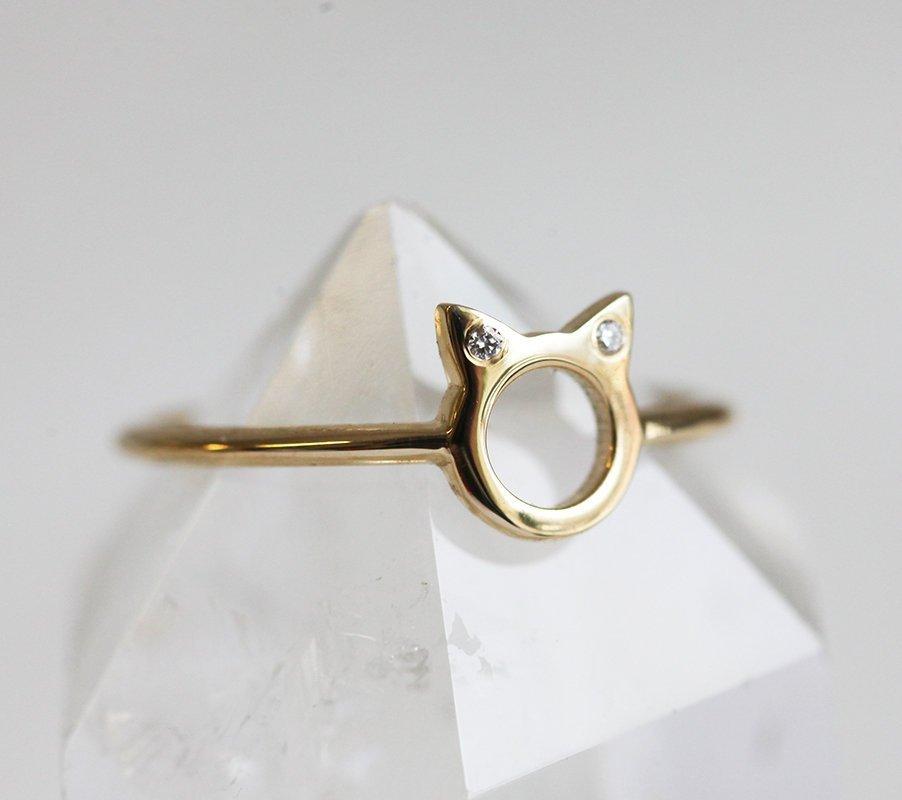A gold ring in the shape of cat ears with round white diamonds on the ears, on a textured background