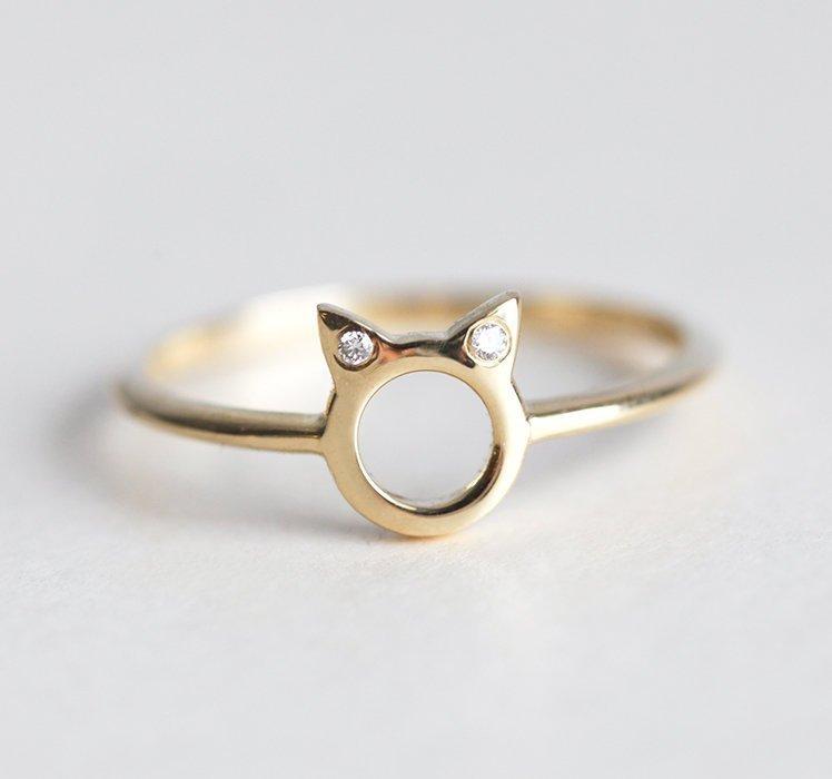 A gold ring in the shape of cat ears with round white diamonds on the ears, on a textured background