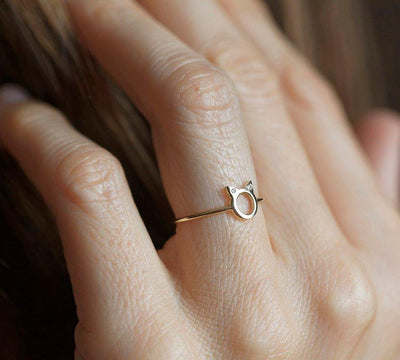 A gold ring in the shape of cat ears with round white diamonds on the ears, on a textured background