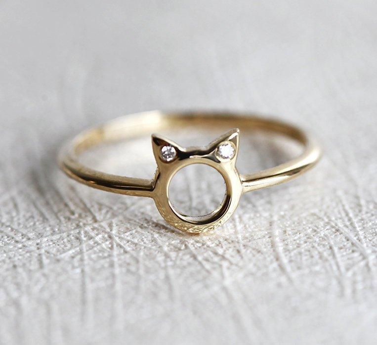 A gold ring in the shape of cat ears with round white diamonds on the ears, on a textured background