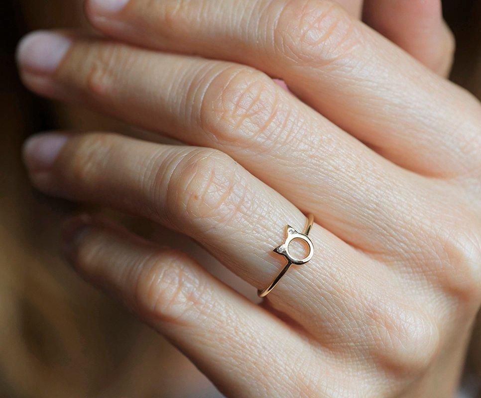 A gold ring in the shape of cat ears with round white diamonds on the ears, on a textured background