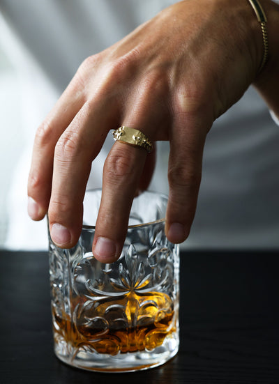 A hand wearing a Snake Gold Signet Ring over a glass of alcohol.