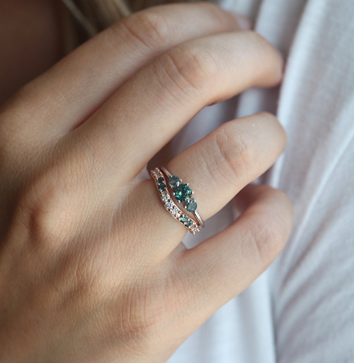 A hand wearing a teal sapphire and moss agate engagement ring. The ring features a central teal mermaid sapphire flanked by round moss agates.