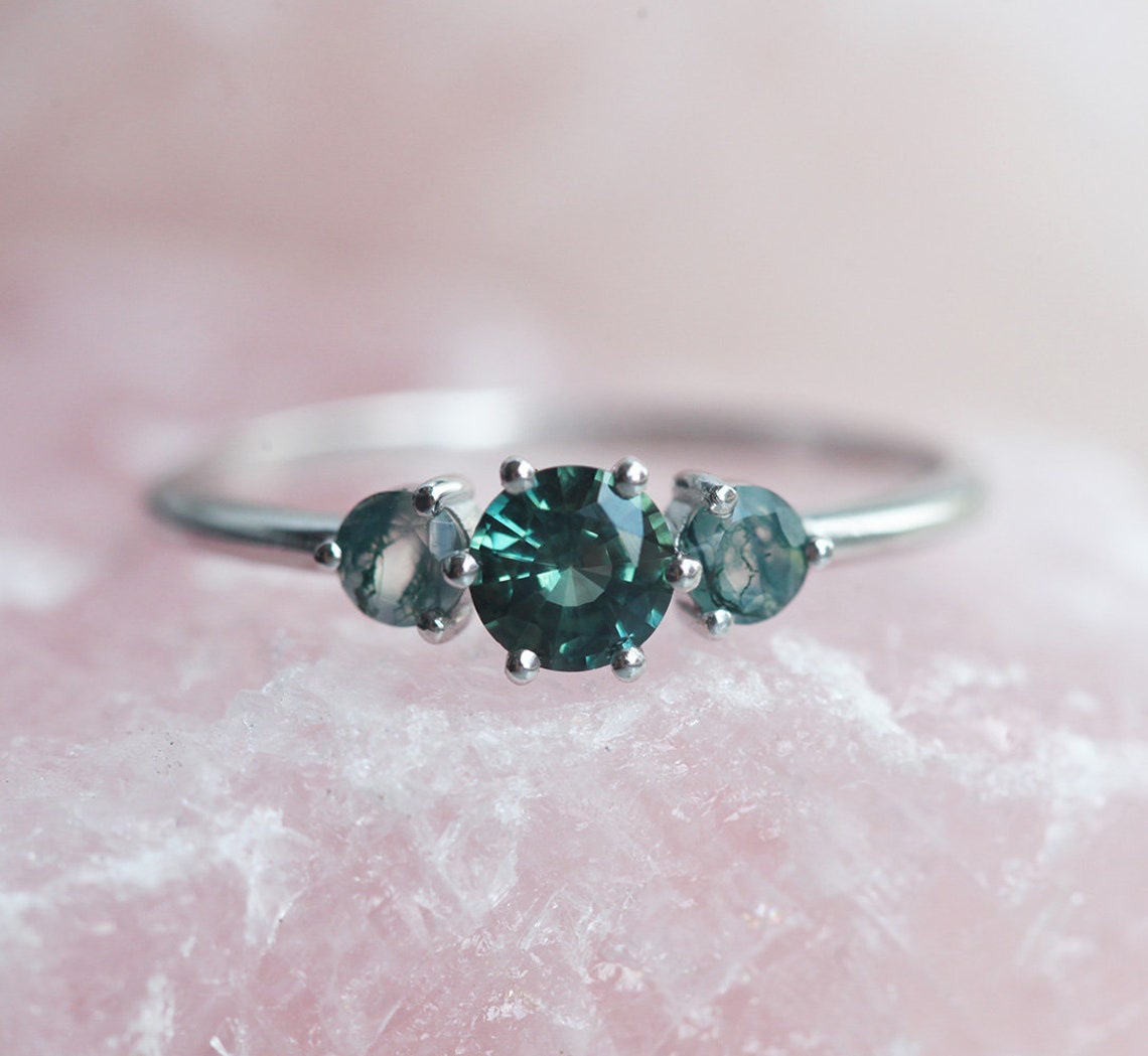 A close-up image of a teal sapphire and moss agate engagement ring. The ring features a central teal mermaid sapphire flanked by two round moss agates, displayed on a pink crystal background.