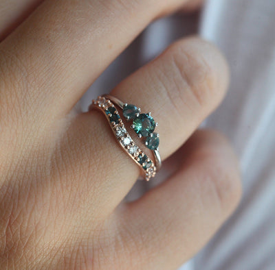 A hand wearing a teal sapphire and moss agate engagement ring, featuring a central teal mermaid sapphire flanked by two round moss agates, paired with a curved band adorned with green gemstones and diamonds.