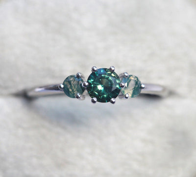 A close-up image of a teal sapphire and moss agate engagement ring. The ring features a central teal mermaid sapphire flanked by two round moss agates, displayed on a light grey background.