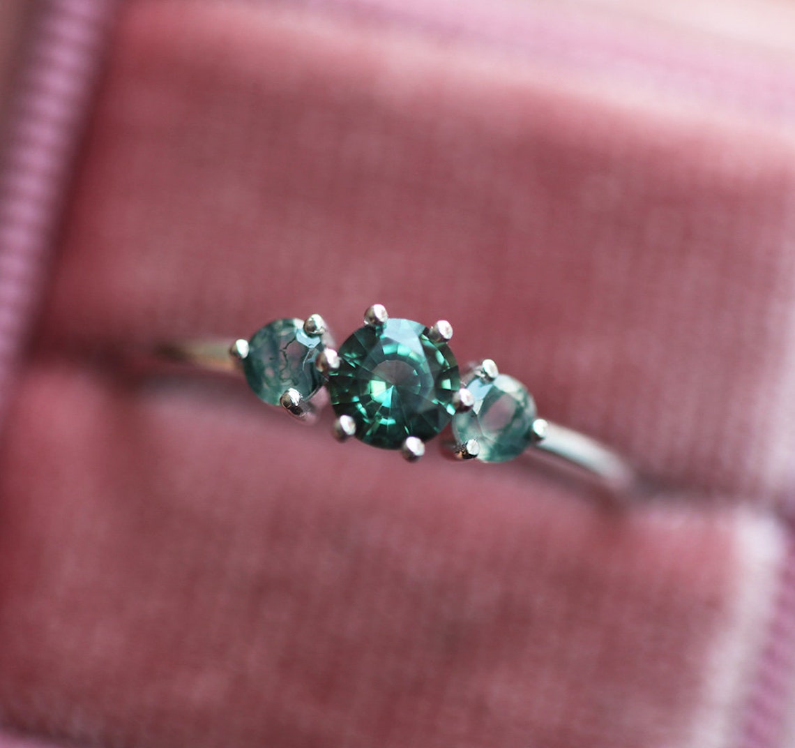 A close-up image of a teal sapphire and moss agate engagement ring. The ring features a central teal mermaid sapphire flanked by two round moss agates, displayed in a pink velvet ring box.