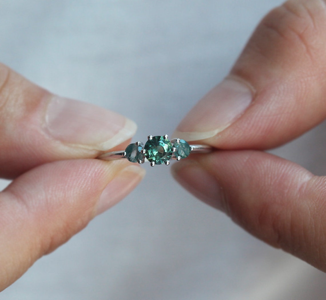 A close-up image of hands holding a teal sapphire and moss agate engagement ring. The ring features a central teal mermaid sapphire flanked by two round moss agates.