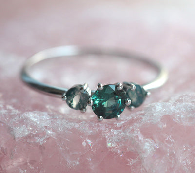 A close-up image of a teal sapphire and moss agate engagement ring. The ring features a central teal mermaid sapphire flanked by two round moss agates, displayed on a pink crystal background.