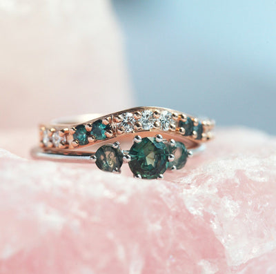 A close-up image of a teal sapphire and moss agate engagement ring, featuring a central teal mermaid sapphire flanked by two round moss agates, paired with a curved band adorned with alternating green gemstones and diamonds. The rings are displayed on a pink crystal background.