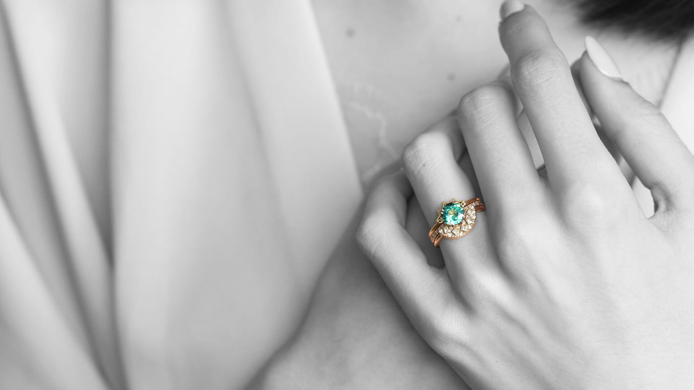 A close-up of a hand wearing a gold ring with a green gemstone and diamonds, set against a soft, blurred background.