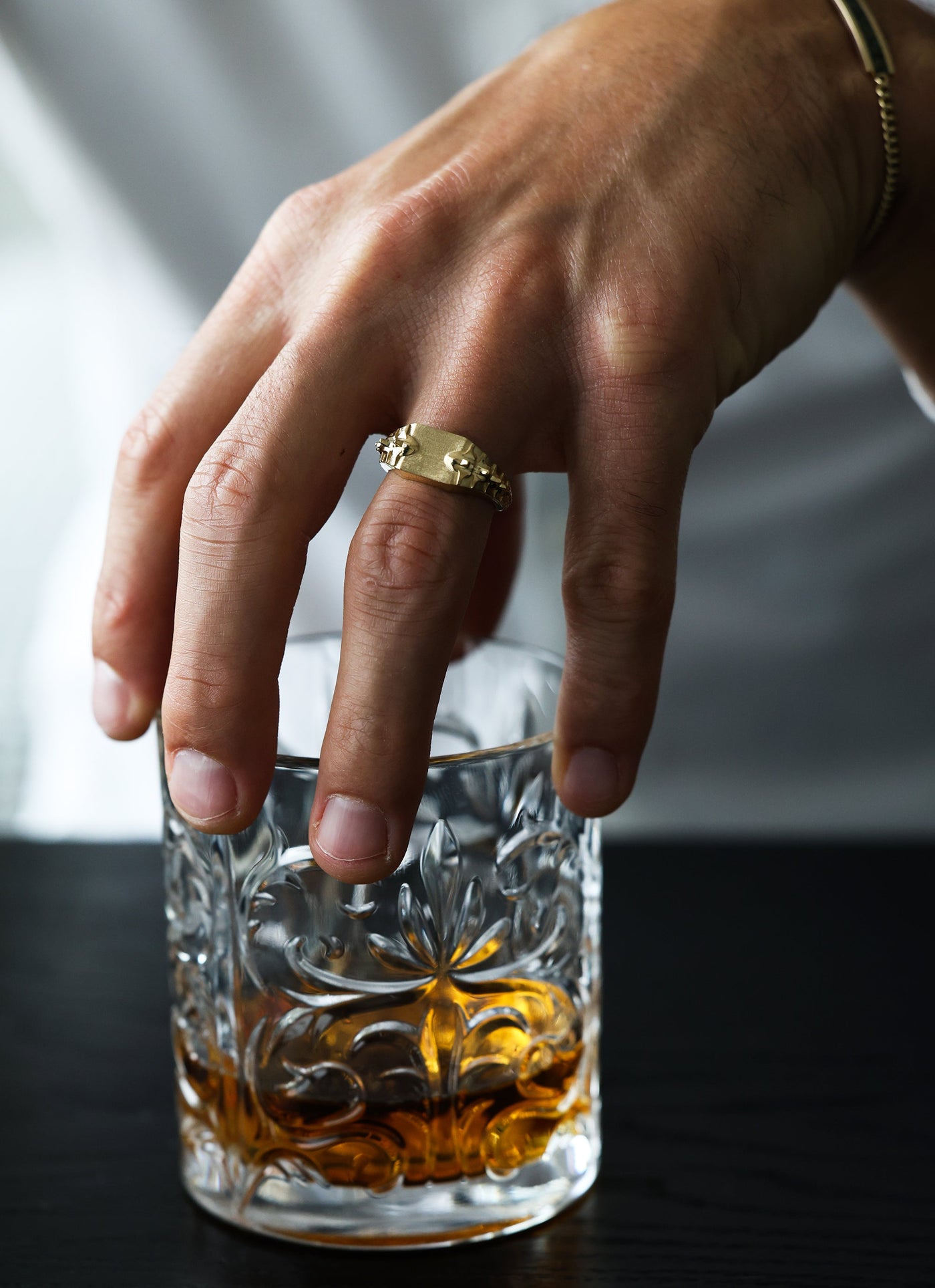 A men's gold signet ring with snake spine detail, held over a glass of alcohol.